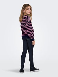 Pihe-puha rózsaszín és sötétkék csíkos pulóver hosszú ujjakkal, sötét farmer szűk szabású nadrággal és fekete magas szárú tornacipővel párosítva.