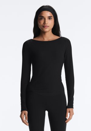 Kvinde iført sort langærmet tætsiddende bluse og sorte leggings, med sort pandebånd, stående mod en ensartet lys baggrund.