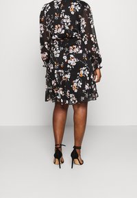 Robe noire à fleurs avec manches longues, ourlet à volants et taille cintrée. Composée d'un tissu transparent avec des motifs de fleurs blanches et orange. Talons hauts noirs.