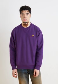 Violetinis megztinis su apvaliu kaklu ir guma apdirbtais rankogaliais, ant krūtinės esantis mažas Carhartt logotipas geltonos ir raudonos spalvos.