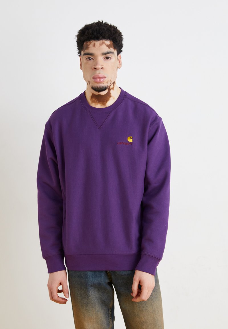 Violetinis megztinis su apvaliu kaklu ir guma apdirbtais rankogaliais, ant krūtinės esantis mažas Carhartt logotipas geltonos ir raudonos spalvos.