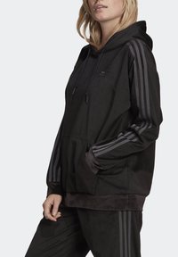 Svart huvtröja med ribbad struktur, med gråa randdetaljer på ärmarna, en framficka och justerbar huva med dragsko.