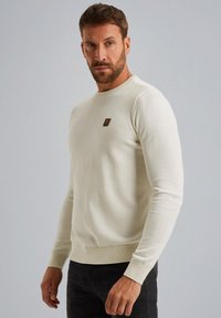 Maglione color crema con una texture a coste, scollatura rotonda e polsini aderenti. Presenta una piccola etichetta logo marrone sul petto.