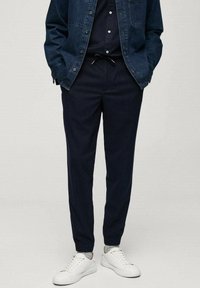 Námořnické modré teplákové kalhoty s elastickým pasem, stahovací šňůrkou, bočními kapsami a zúženými nohavicemi, doplněné bílými teniskami a tmavým džínovým sáčkem.