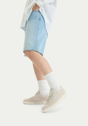 Persona che indossa pantaloncini di jeans color azzurro chiaro, calze bianche, sneaker beige e una maglietta bianca con la mano in tasca, in piedi su uno sfondo bianco.