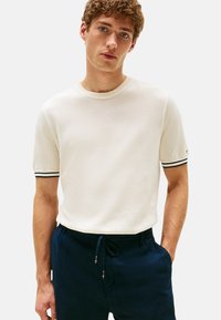 Maglietta bianca a maniche corte con scollo a girocollo, dettagli a righe blu navy e bianche sulle maniche, indossata con pantaloni navy con coulisse.