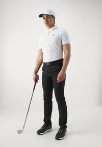 Vit pikétröja med en svart logotyp, svarta byxor och svarta golfskor, håller en golfklubba i en neutral miljö.