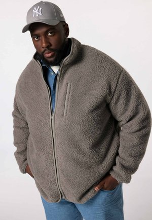 Grijze fleece jas met een rits aan de voorkant, zachte textuur en zijzak. Gedragen over een denim shirt, gecombineerd met blauwe jeans en een grijze pet.
