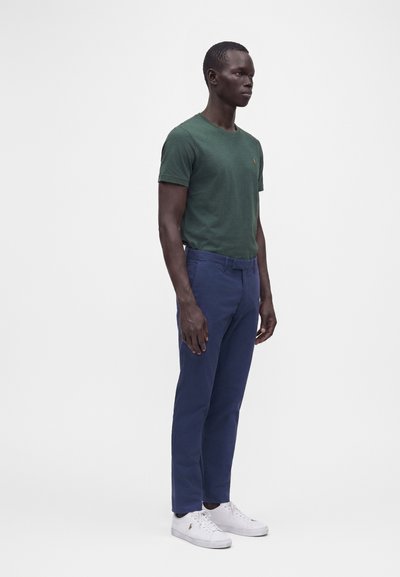 Grand homme debout portant une chemise verte à manches courtes, un pantalon bleu et des baskets blanches sur un fond clair uni.