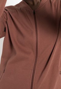 Brun zip-jacka i mjukt tyg, med hög krage och sidofickor, med en matt yta och minimalistisk design.