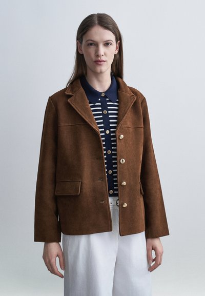 Jeune femme portant une veste marron boutonnée sur une chemise rayée bleu marine et blanche, associée à un pantalon blanc, debout devant un fond uni.