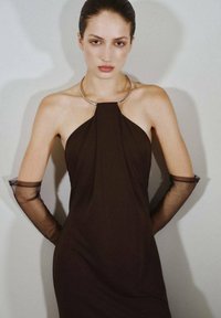 Brązowa sukienka na ramiączkach o strukturalnym fasonie, z metalowym kołnierzem w kolorze srebrnym oraz przezroczystymi, długimi czarnymi rękawiczkami z elastycznymi mankietami.