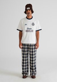 Man wearing brown beanie, white T-shirt with "Escape Berlin" text, black and white plaid pants, and brown shoes standing against plain background.