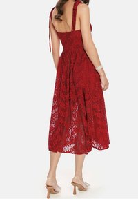 Abito midi rosso in pizzo con spalline regolabili, corpetto aderente e gonna fluida. Presenta un motivo floreale in pizzo e una texture trasparente. Indossato con tacchi trasparenti.