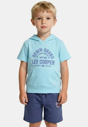 Jeune garçon aux cheveux blonds courts portant un t-shirt à capuche bleu clair et un short bleu marine, debout face à l'avant.
