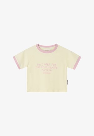 Krém rövid ujjú póló rózsaszín szegéllyel és rózsaszín felirattal az elején, amely így szól: "Csak a Pilates utáni matcháért vagyok itt", címke az oldalvarráson.