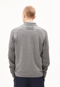 Pull en tricot gris avec un col, présentant des manches et un ourlet côtelés. Patch rectangulaire notable au niveau du col. Texture lisse.