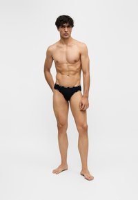 Jeune homme debout pieds nus portant un slip noir avec un texte blanc sur la ceinture sur un fond uni clair.