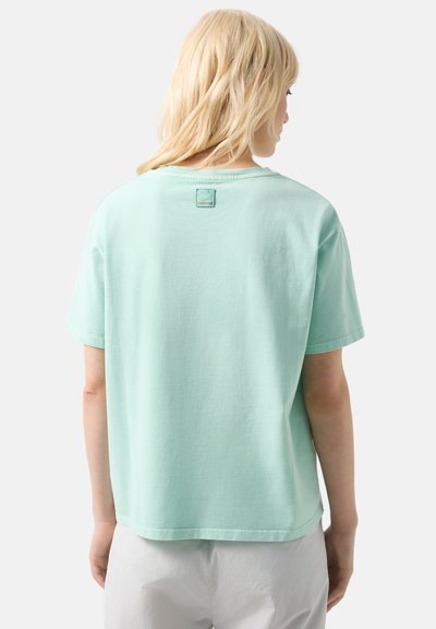 Blonde vrouw van achteren, gekleed in een loszittend mintgroen T-shirt met korte mouwen en een witte broek, tegen een effen achtergrond.