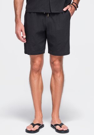 Hombre con pantalones cortos negros y chanclas negras, de pie con una mano en el bolsillo y una pulsera de cuentas en la muñeca.