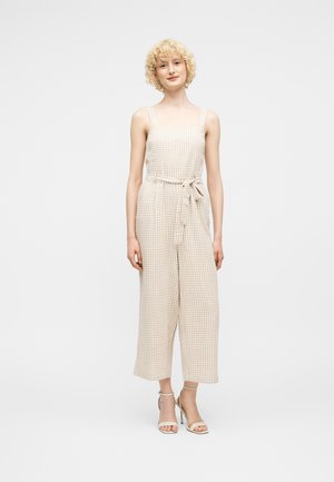 Junge Frau mit lockigem blondem Haar, die einen ärmellosen Jumpsuit in Beige und Weiß mit Karo-Muster und gebundenem Taillengürtel sowie beige Sandaletten mit hohen Absätzen trägt.