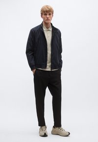 Tumesinine lukuga jope helebeeži pologa, seljas mustad püksid ja beežid tossud, lihtsa valge taustaga.