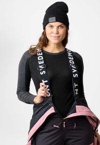 Berretto di lana nera, maglietta a maniche lunghe grigia e salopette nera con dettagli rosa e spalline regolabili. Tasche laterali con zip visibili.