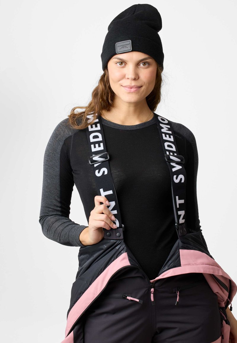 Berretto di lana nera, maglietta a maniche lunghe grigia e salopette nera con dettagli rosa e spalline regolabili. Tasche laterali con zip visibili.