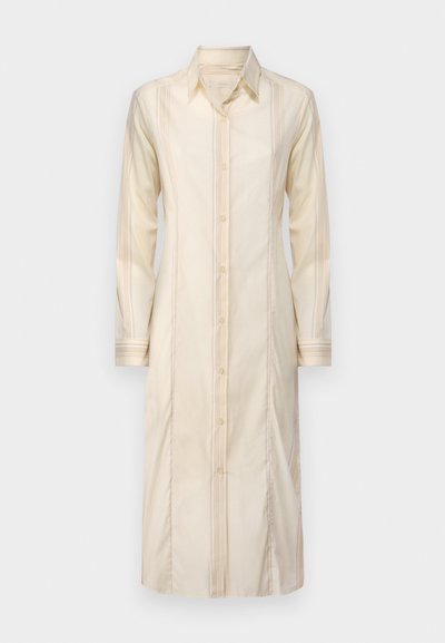 Rochie lungă din material crem, cu guler, închidere cu nasturi pe față și dungi verticale; are mâneci lungi și o siluetă dreaptă.
