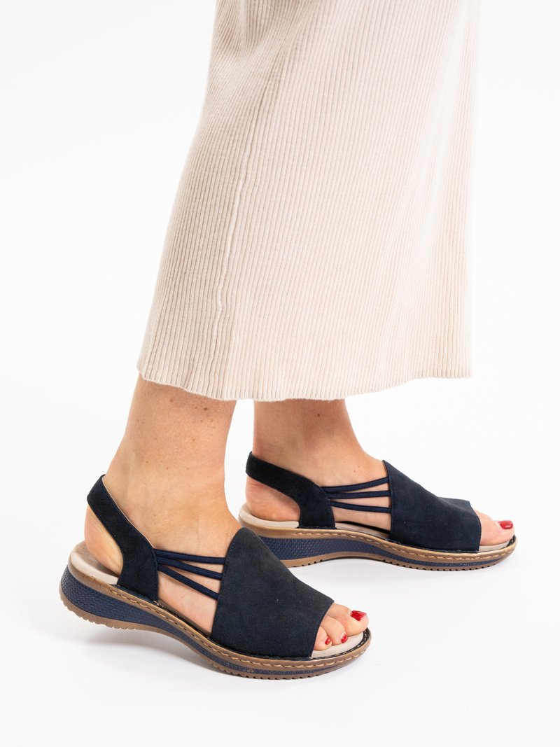 Pies con sandalias negras de punta abierta y tiras elásticas, con esmalte de uñas rojo, combinados con una falda beige de punto acanalado que llega hasta la mitad de la pantorrilla.