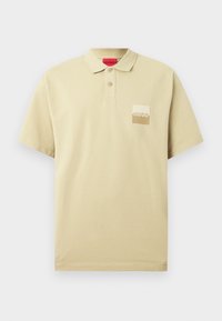 Polo à manches courtes beige avec deux boutons et un petit patch carré en logo portant la mention "HUGO" sur la poitrine gauche.
