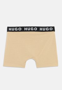 Šviesiai rudos spalvos bokso tipo apatiniai šortai su minkšta tekstūra, su juodu elastiniu diržu, kuriame kartojasi baltas "HUGO" logotipas. Vidutinio ilgio kirpimas su siūlėmis.