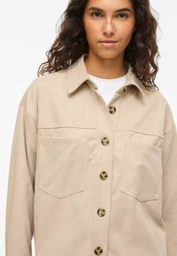 Bēša pogājama krekla krekls no teksturētas auduma, ar lielām kabatām, smailu apkakli un zelta toņa pogām priekšpusē.