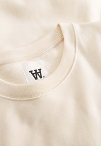 Sweat-shirt beige à col rond avec un col côtelé, une texture douce et une étiquette avec des lettres noires. Le matériau est lisse et décontracté.