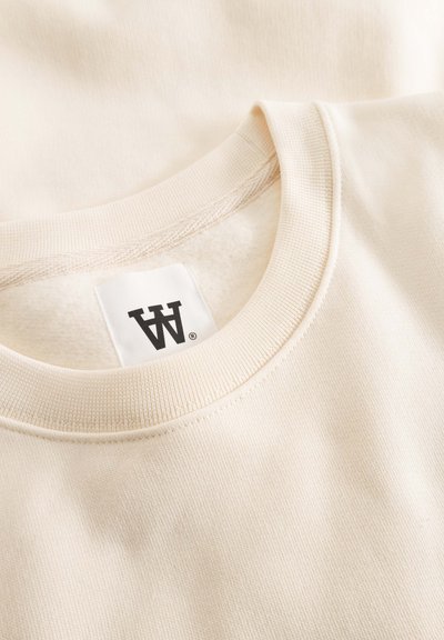 Sweat-shirt beige à col rond avec un col côtelé, une texture douce et une étiquette avec des lettres noires. Le matériau est lisse et décontracté.