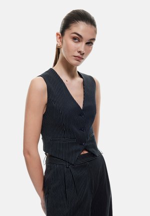 Gilet à rayures marines avec un décolleté en V profond, sans manches, cinq boutons avant et coupe ajustée. Tissu texturé avec une forme structurée.