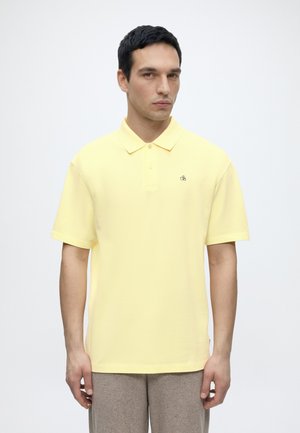 Homme portant un polo jaune pâle à manches courtes et un pantalon beige, debout devant un fond blanc uni.