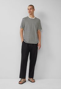 T-shirt à manches courtes rayé en crème et bleu marine, associé à un pantalon noir ample et des sandales marron. Design simple et coupe décontractée.
