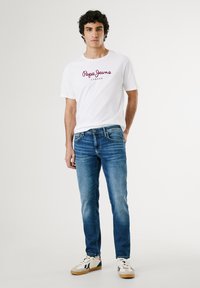 Camiseta blanca con el logotipo rojo "Pepe Jeans London", combinada con unos vaqueros de mezclilla azules y deportivas blancas con detalles en negro y rojo.