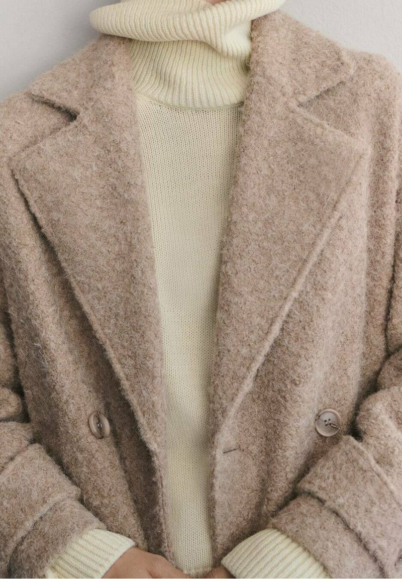Personne portant un manteau en laine beige duveteux sur un pull à col roulé côtelé crème, les mains partiellement visibles au niveau de la taille.