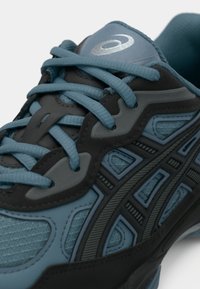 Sportskor i teal och svart, med andningsbart mesh, mönstrade överdrag och platta snören. Accenter inkluderar en reflekterande logotyp på tungan.