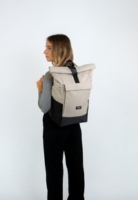 Sac à dos à rabat en beige et noir, fabriqué en tissu durable. Dispose d'une poche zippée à l'avant et d'une sangle noire avec une boucle.