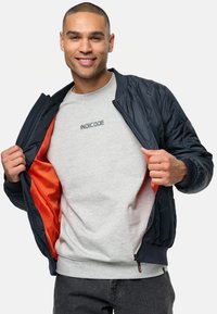 Chaqueta bomber acolchada en navy con un forro naranja, que presenta un cierre con cremallera y puños acanalados. Sudadera gris por debajo con el logo "INDICODE".