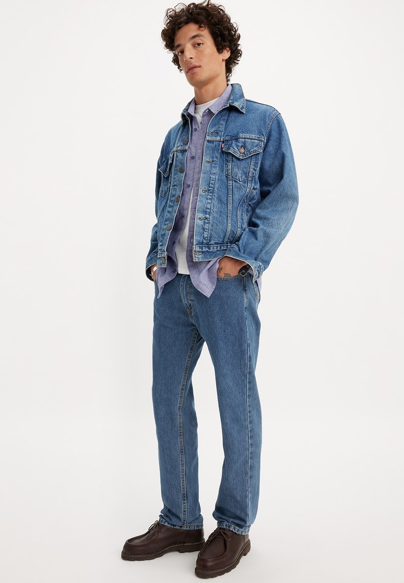 Veste en denim et jeans de couleur bleu clair, superposés à une chemise légère violette. Chaussures marron. Design décontracté avec une texture en denim classique et un matériel traditionnel.