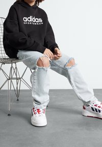 Musta huppari, jossa valkoinen "adidas"-logo, kuluneet vaaleansiniset farkut revityillä polvilla ja valkoiset lenkkarit, joissa on punaisia ja sinisiä yksityiskohtia.