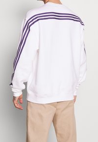 Vit sweatshirt med lila ränder över axlarna och ner längs ärmarna, med en avslappnad passform och ribbade ärmslut och fåll.