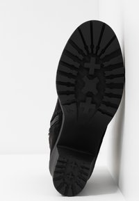 Semelle d'une botte noire avec des motifs de crampons profonds et des croix, inclinée contre une surface blanche en coin.