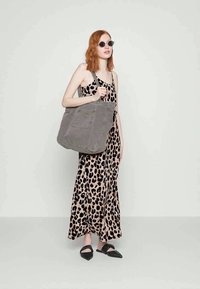Frau mit roten Haaren, die schwarze Sonnenbrille und ein Maxikleid mit Leopardenmuster trägt, eine große graue Tragetasche und schwarze spitze Ballerinas hält.