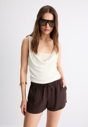 Femme aux longs cheveux bruns porte de grandes lunettes de soleil sombres, un haut blanc sans manches drapé, un short brun foncé et des bracelets dorés à un poignet.