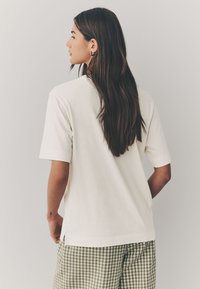 Maglietta a maniche corte color crema con vestibilità ampia, spacchi laterali e texture liscia, abbinata a pantaloncini verdi a quadri.